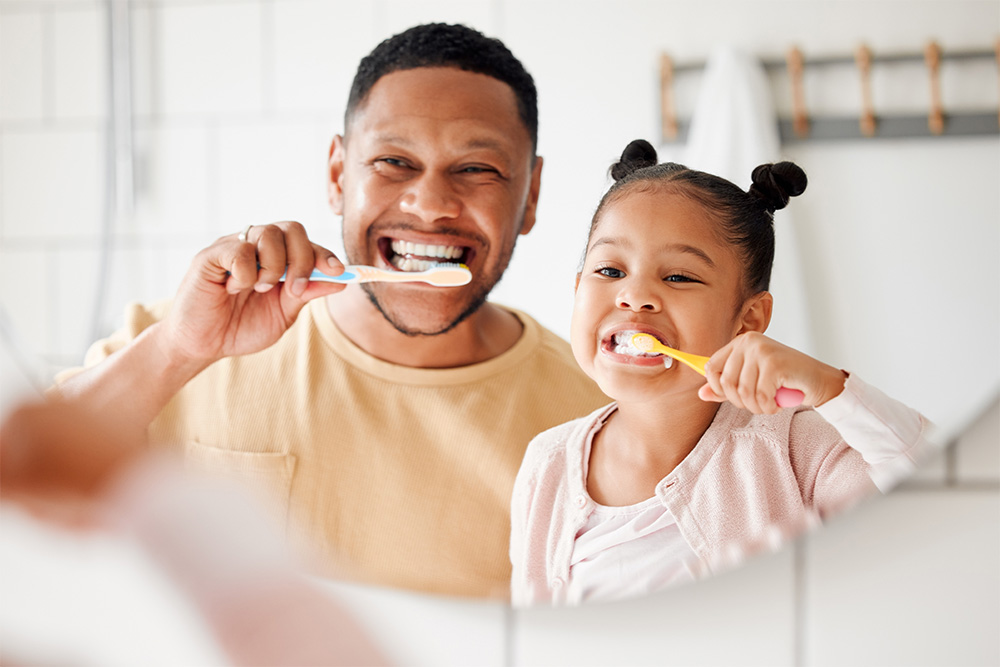 A man and a young girl happily brushing their teeth together in a bright bathroom setting.